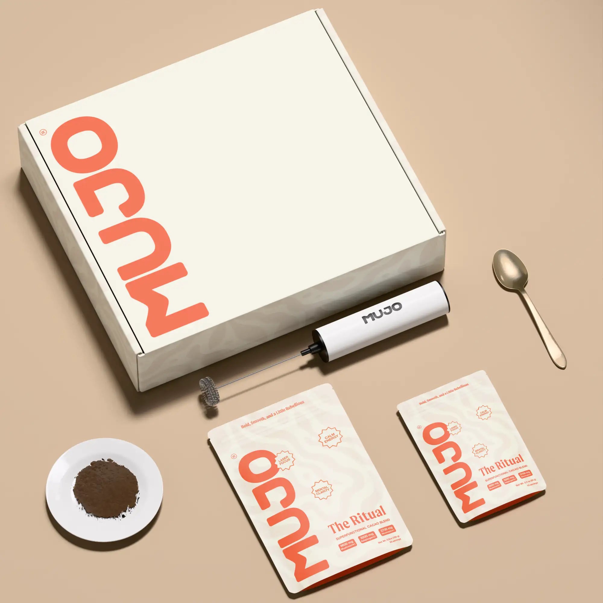 White cardboard box with orange ’ocum’ branding and black trim, containing a mujo handheld frother, two packets of ocum the ritual powder, a small white dish holding brown powder, and a metallic spoon.